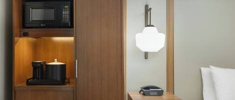 A hotel room with a microwave, a coffee maker, a bedside table with a lamp, and a bed.