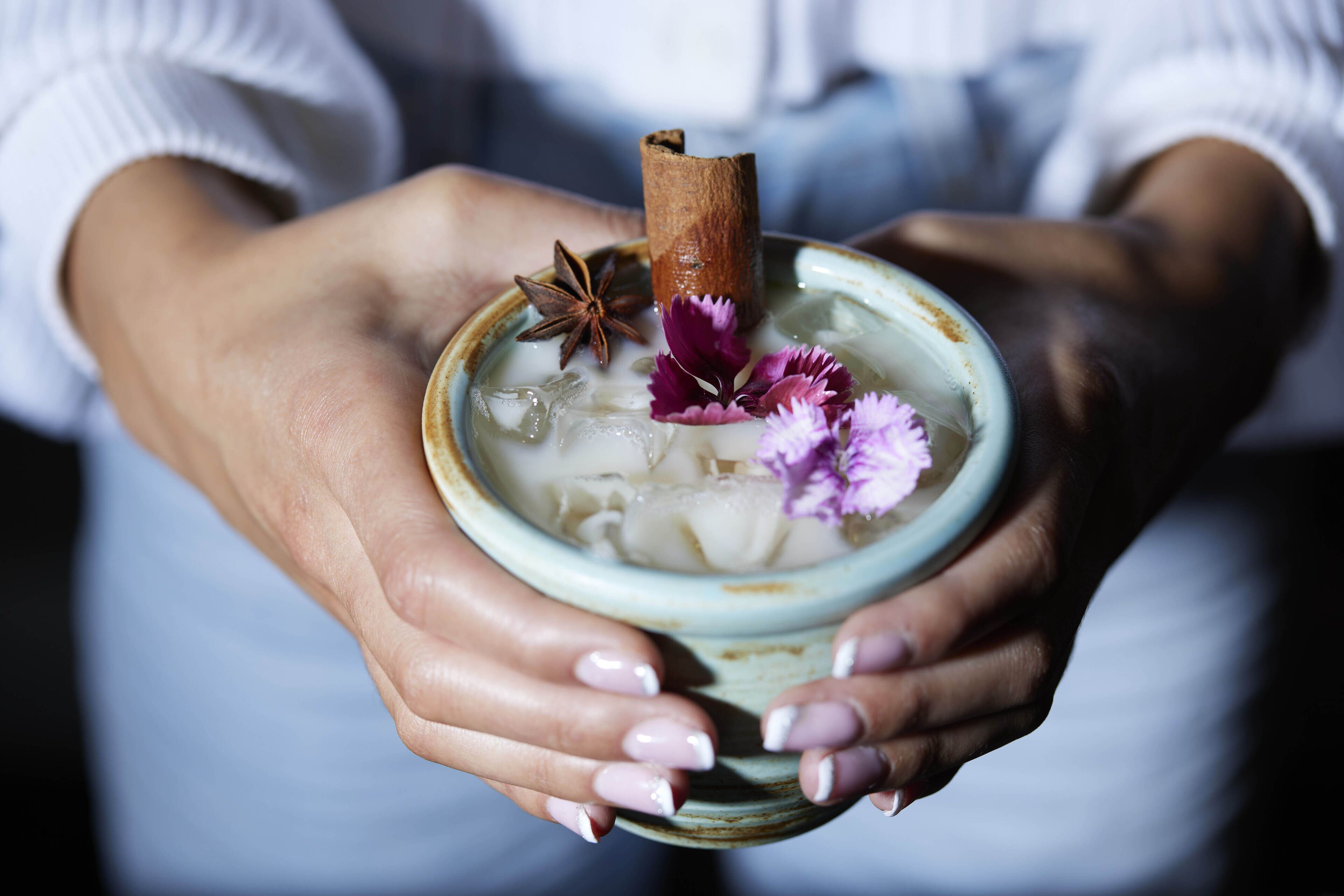 A person holding a ceramic bowl with a beverage, decorated with a cinnamon stick and purple flowers.