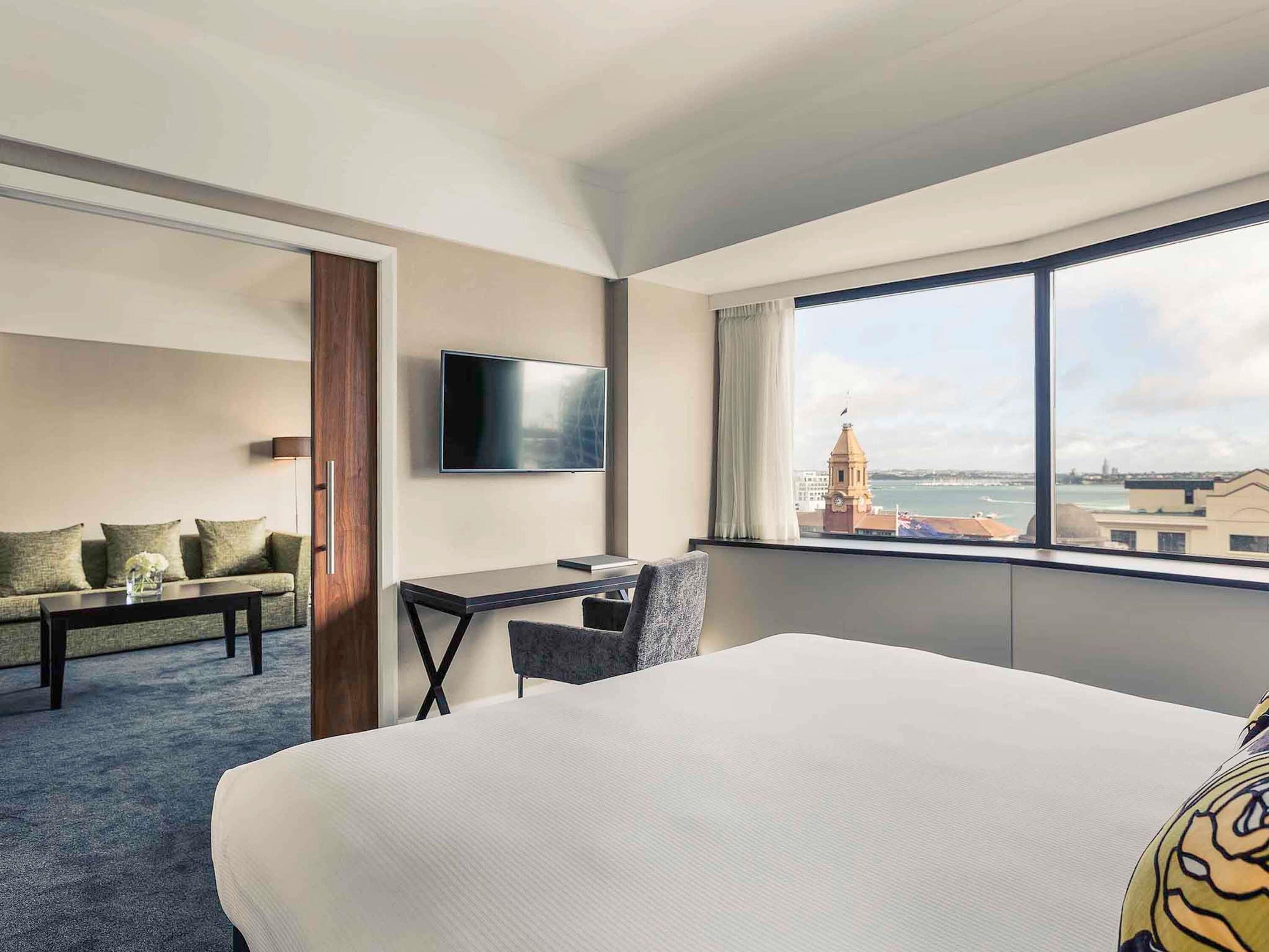 A hotel room with a bed, a desk, a chair, a sofa, a TV, and a large window with a view of a tower and water.
