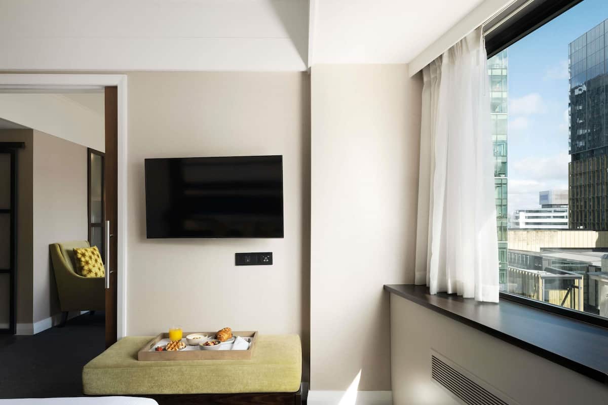 A hotel room with a bed, a small seating area with a tray of breakfast, a flat-screen TV mounted on the wall, and a window with a city view.
