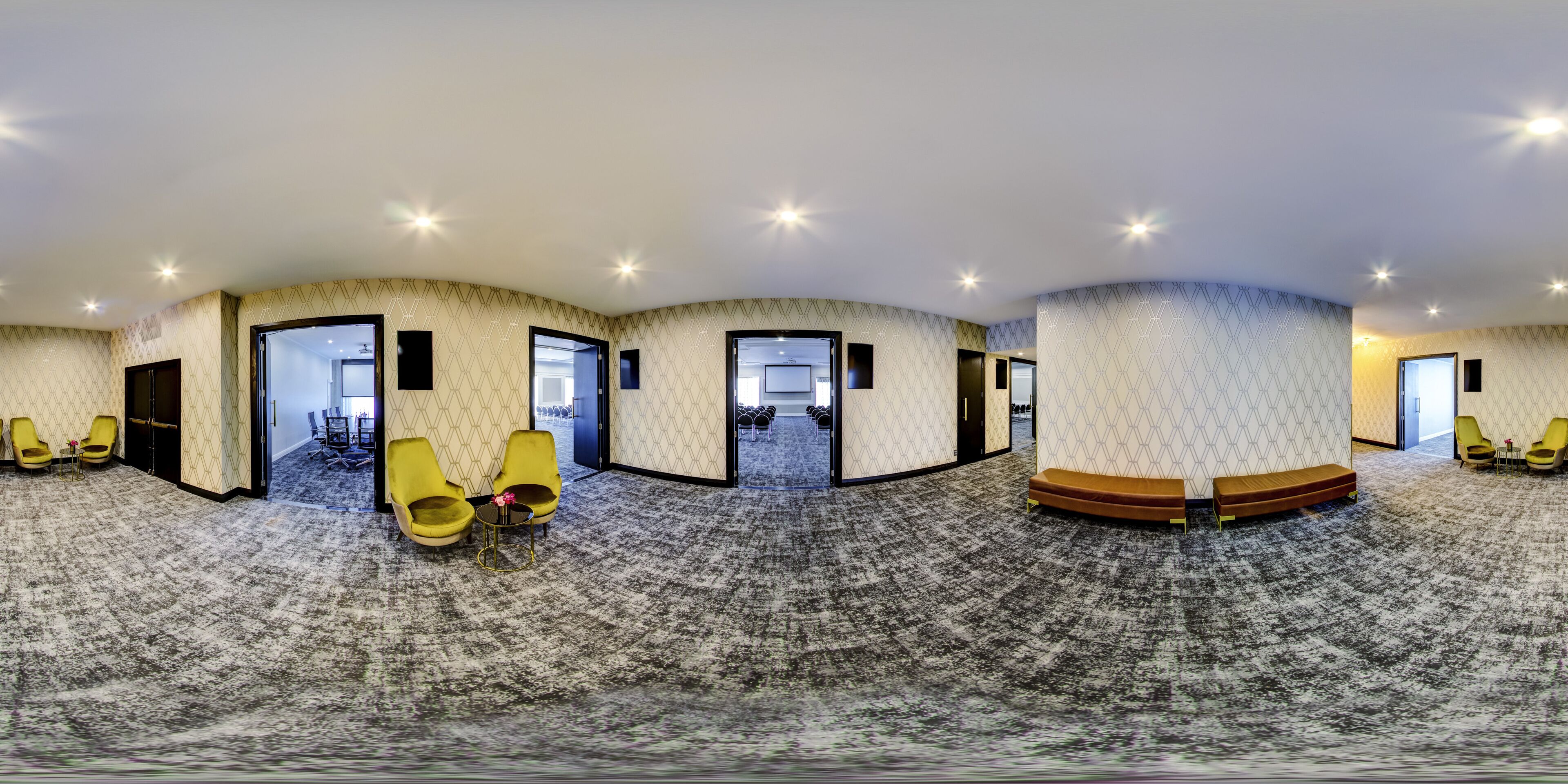 A panoramic view of a hotel lobby with a carpeted floor, yellow chairs, and a seating area with a bench.