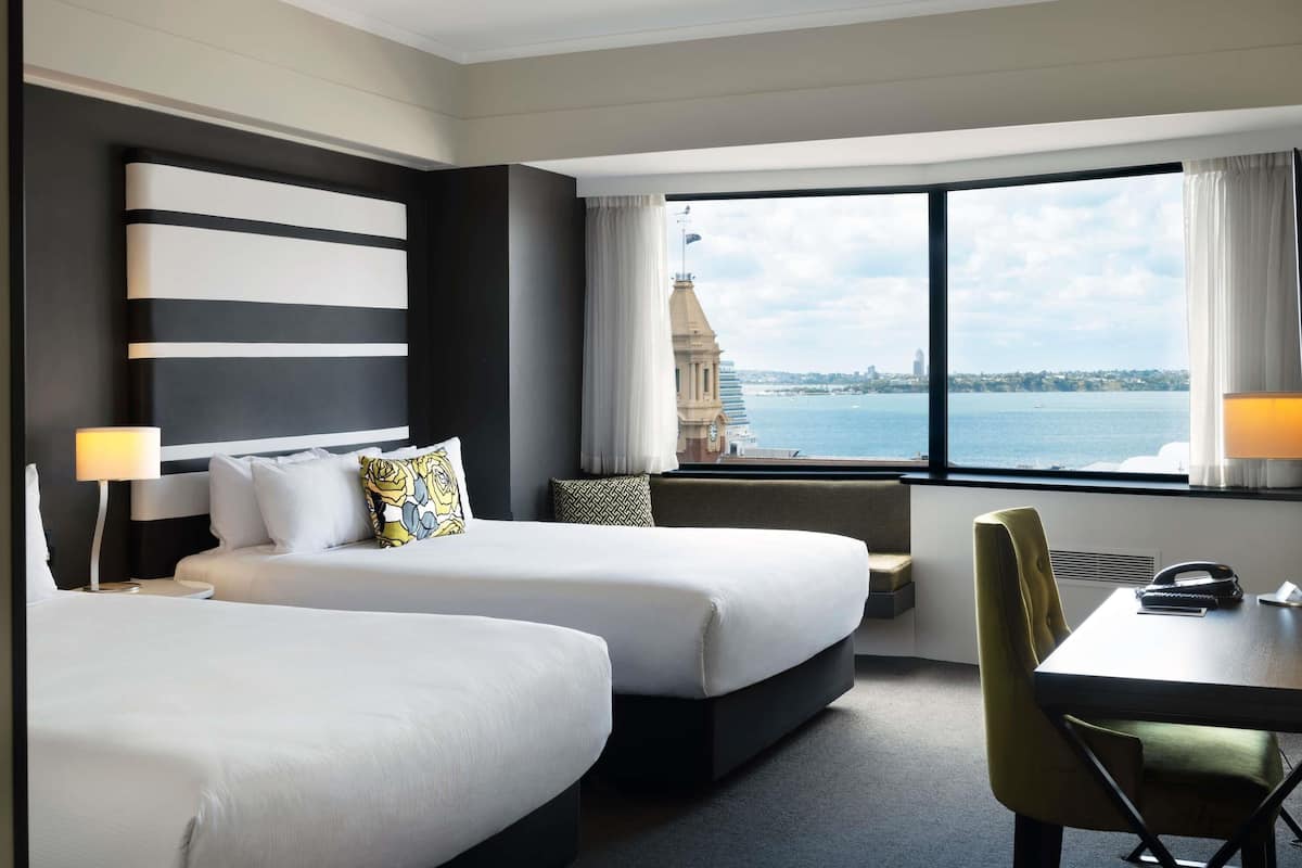 A hotel room with two beds, a desk, a chair, a lamp, and a view of a body of water and a distant building.