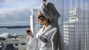 Eine Person in einem weißen Bademantel hält ein Glas mit einem Getränk und steht an einem Fenster mit Blick auf Gebäude und Wasser.