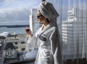 A person in a white bathrobe holding a glass of beverage, standing by a window with a view of buildings and water. - Mövenpick Hotel Auckland (Auckland)