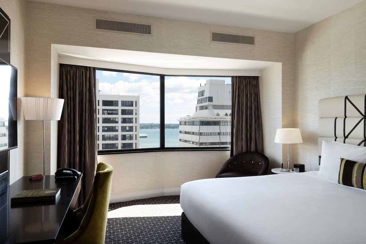A hotel room with a large bed, a desk, a chair, a TV, and a view of a cityscape through a large window.
