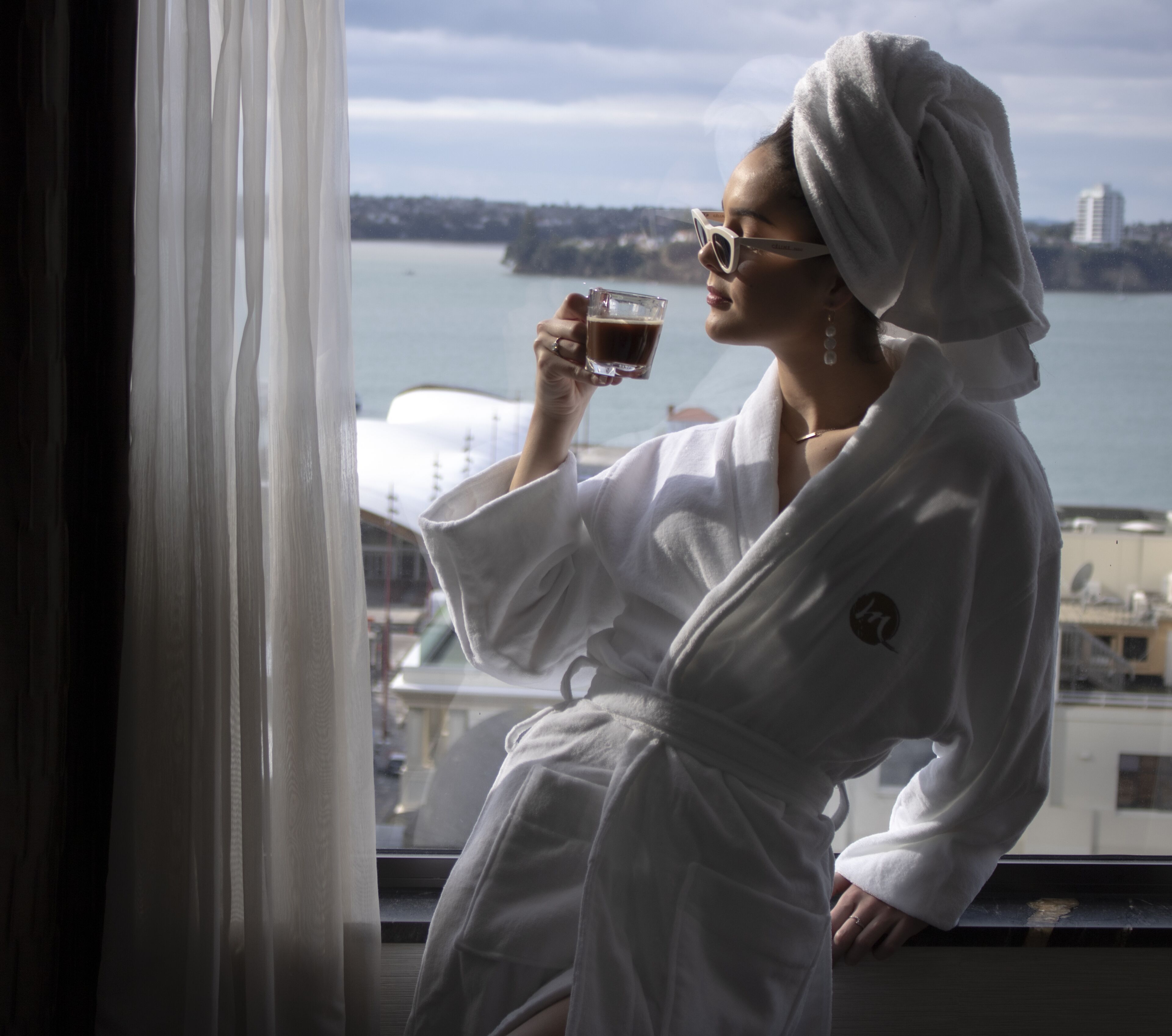 A person in a white bathrobe sitting by a window with a view of a body of water and buildings.