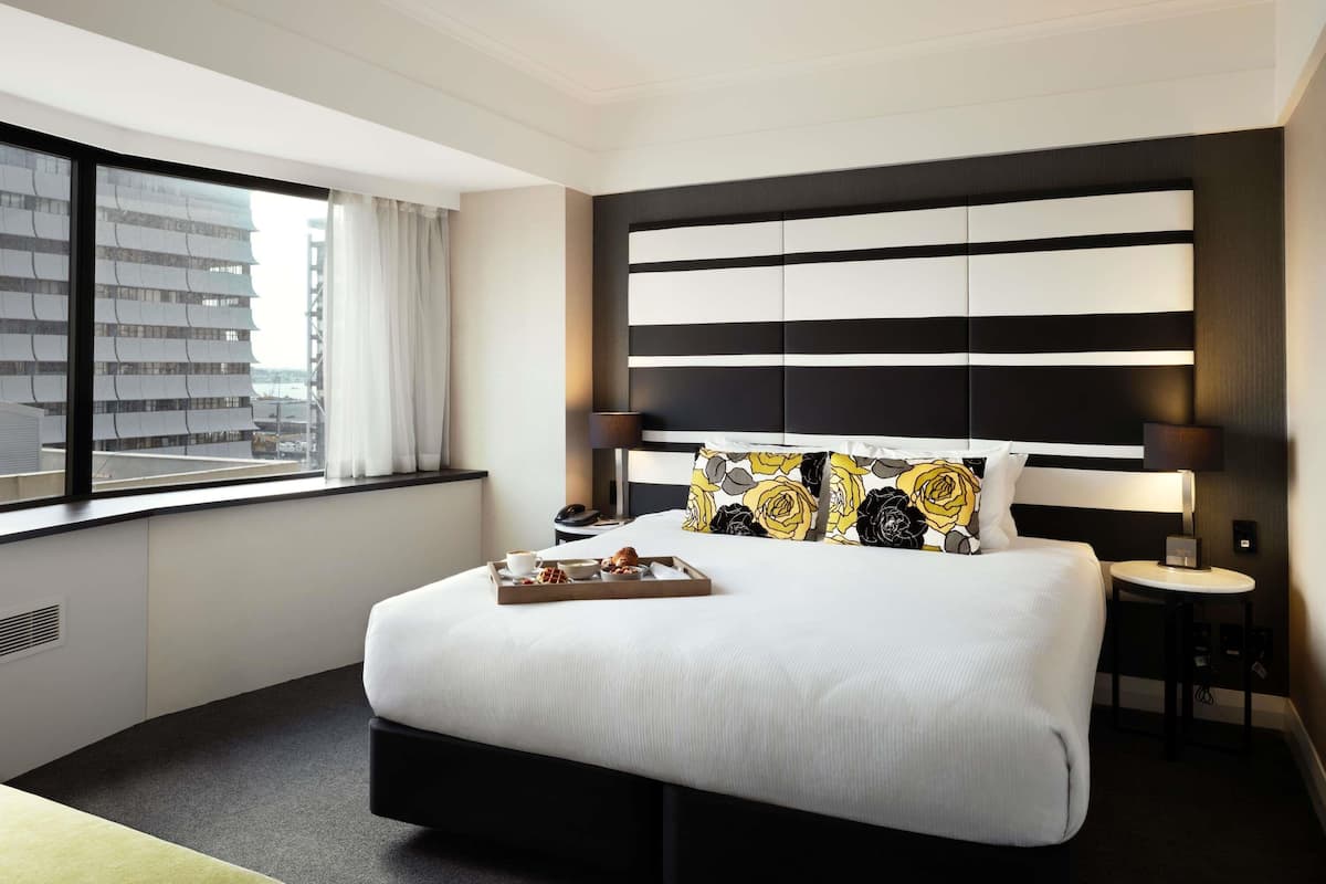 A modern hotel room with a large bed, a bedside table, a lamp, and a view of a building outside.