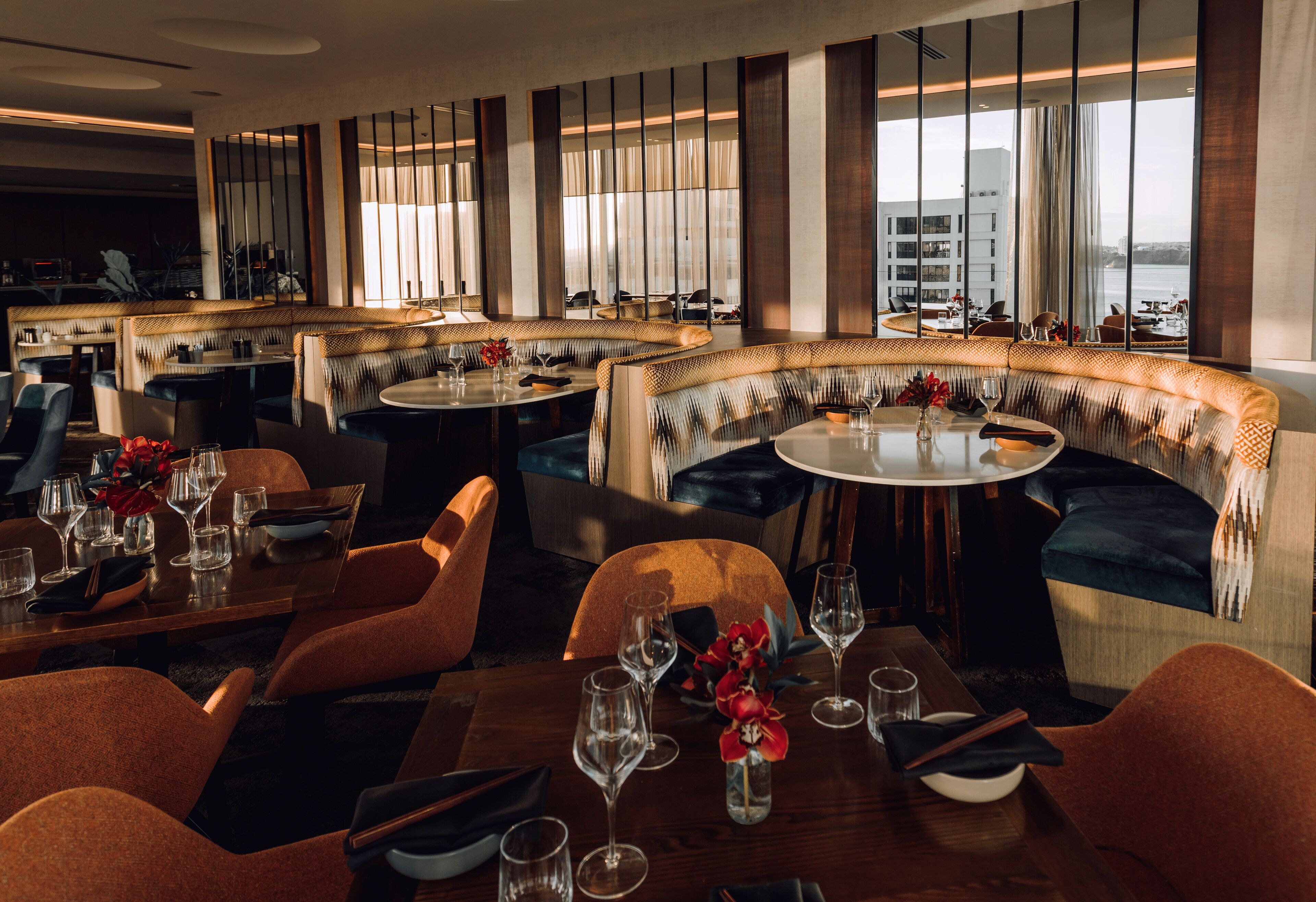 A modern restaurant with round tables and cushioned seating, large windows with curtains, and a view of the city.
