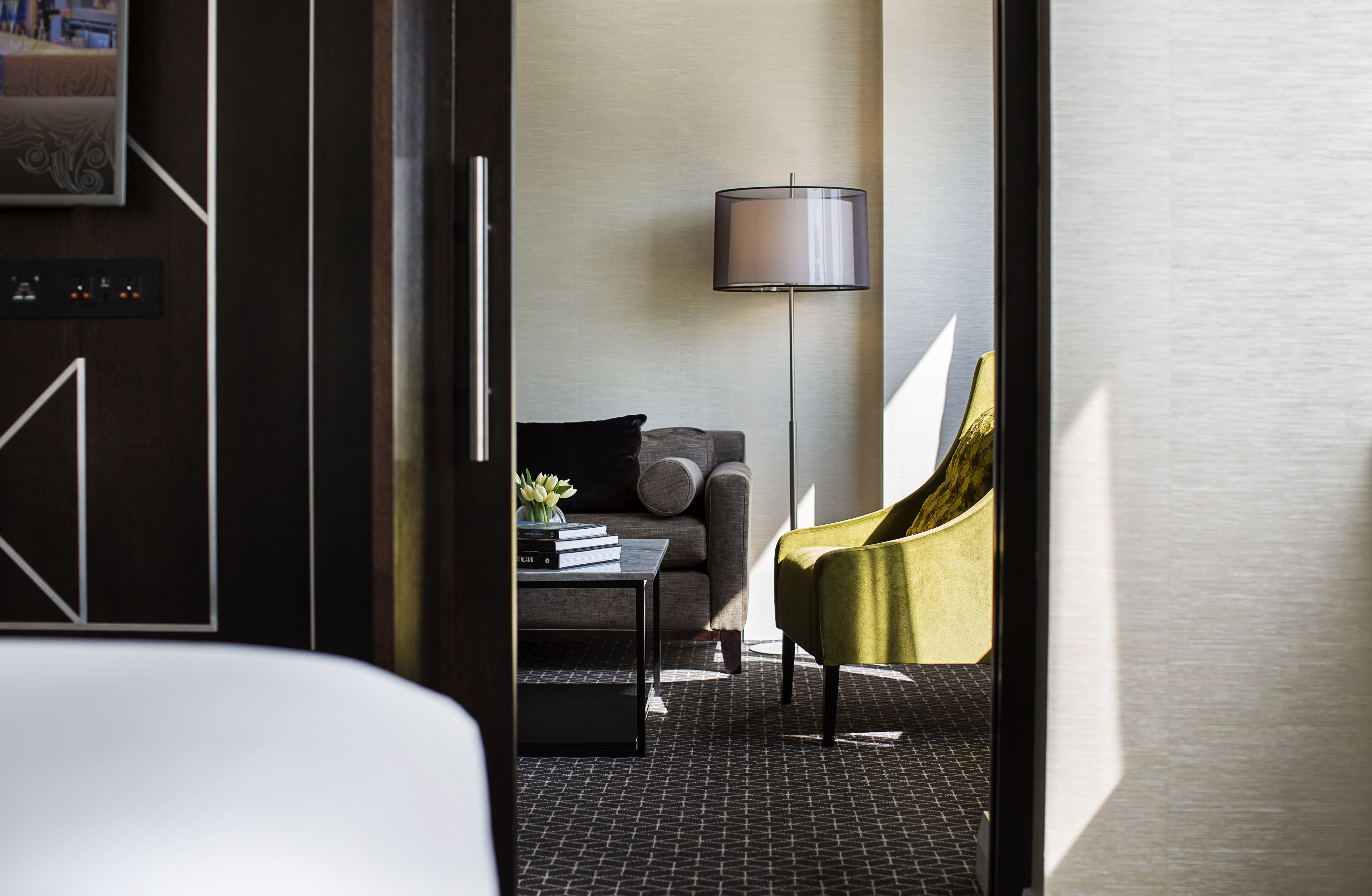 A modern hotel room with a dark wood floor, a grey sofa, a small table with books, a floor lamp, and a yellow armchair.