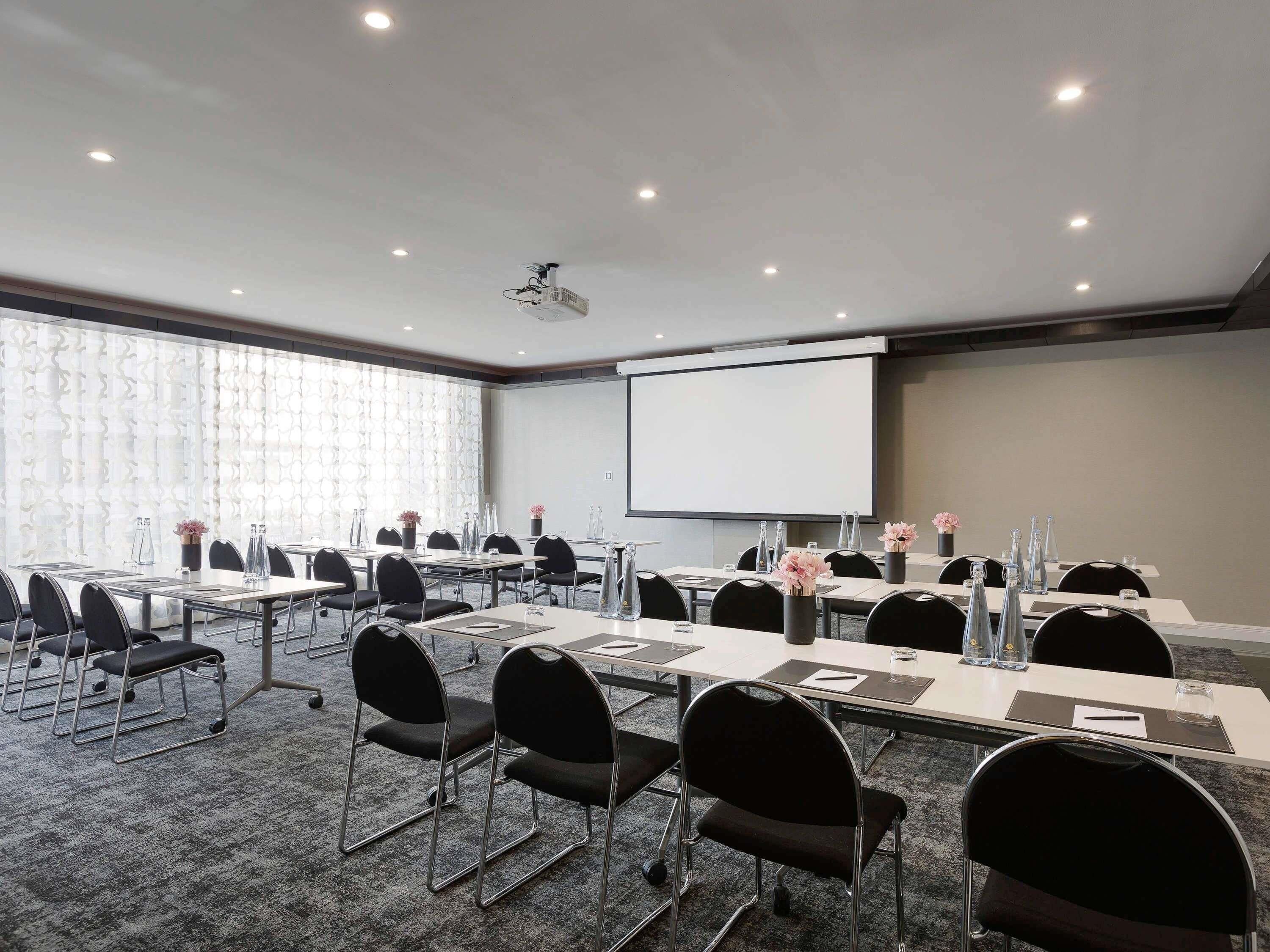 Une salle de conférence équipée de tables, de chaises, d’un écran de projection et d’une grande fenêtre.