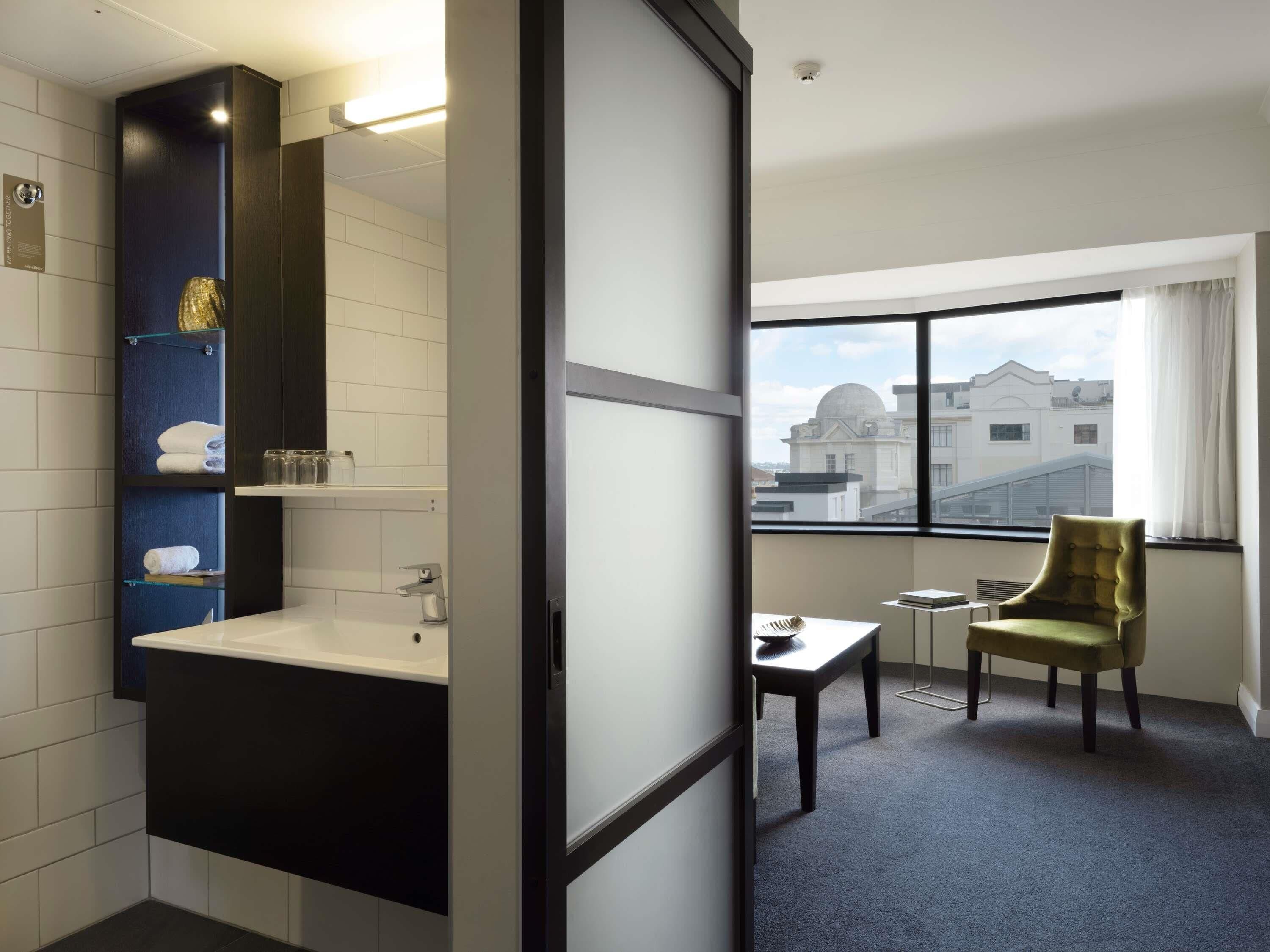 A hotel room with a modern bathroom, a small table with a chair, and a view of buildings through the window.
