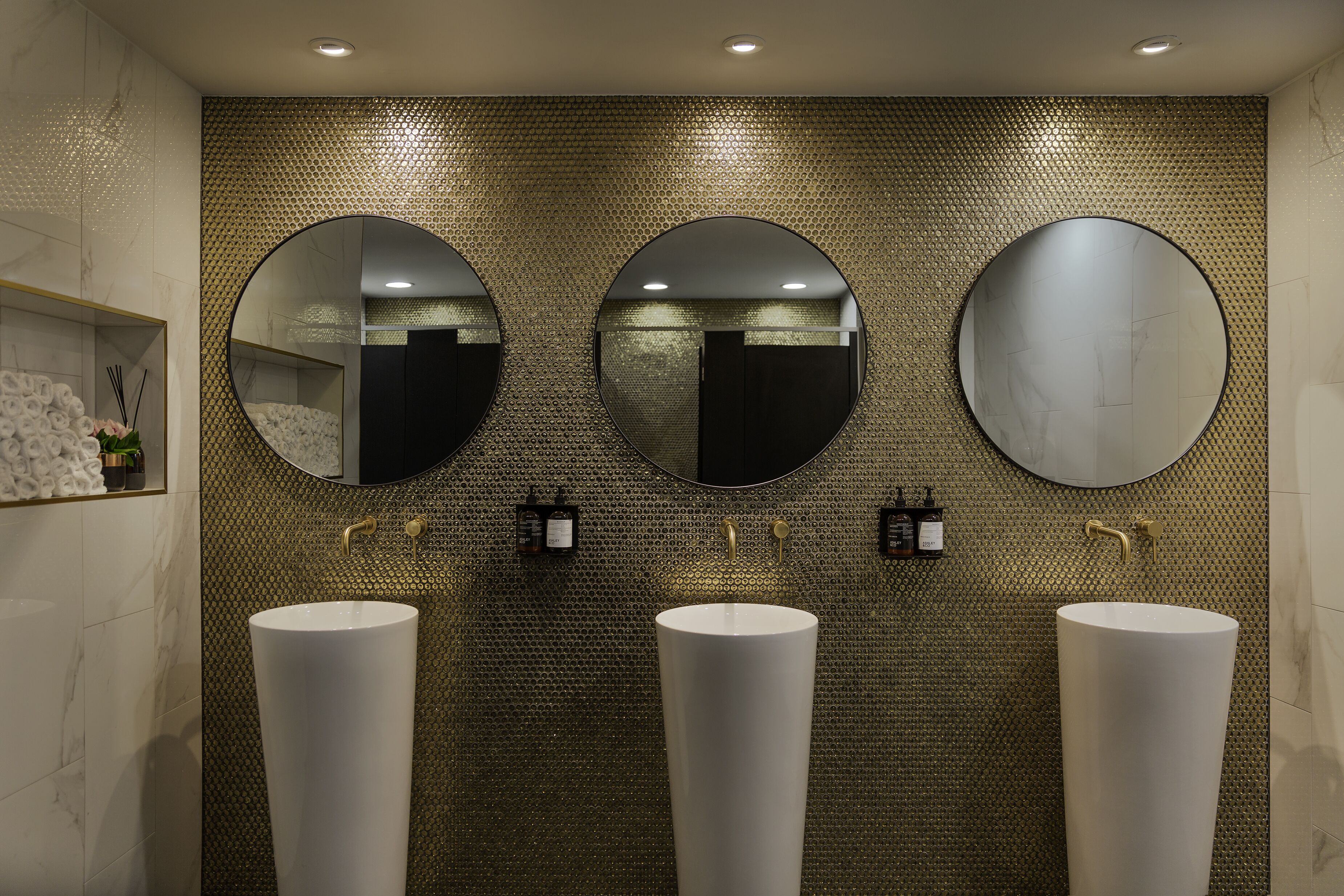 A bathroom with three sinks, each with a large mirror above. The wall has a textured pattern, and there are two small bottles on the wall.