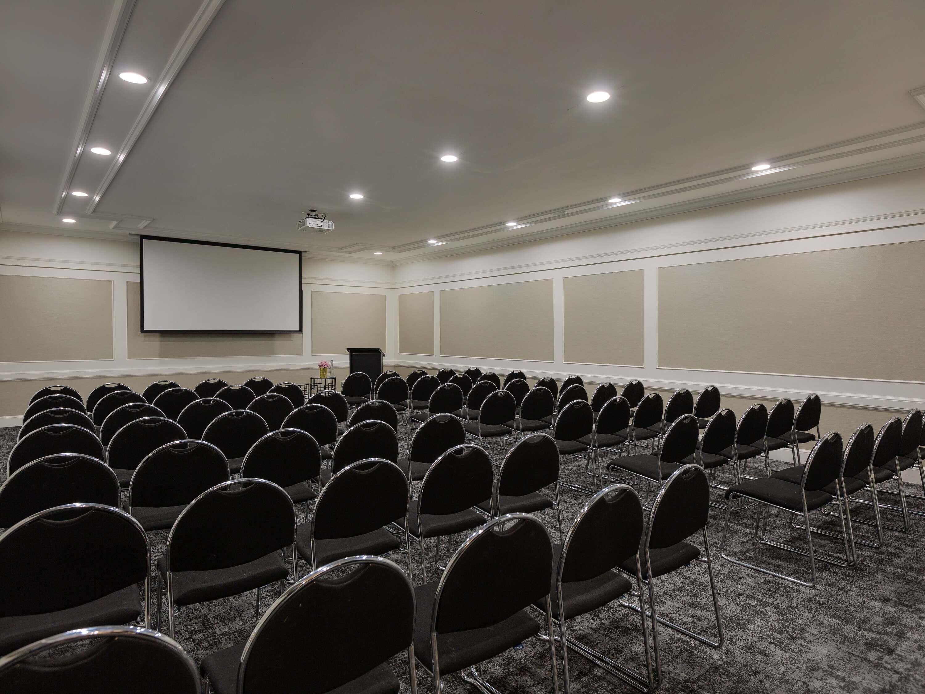 Une salle de conférence avec des rangées de chaises, un écran de projection et un pupitre.