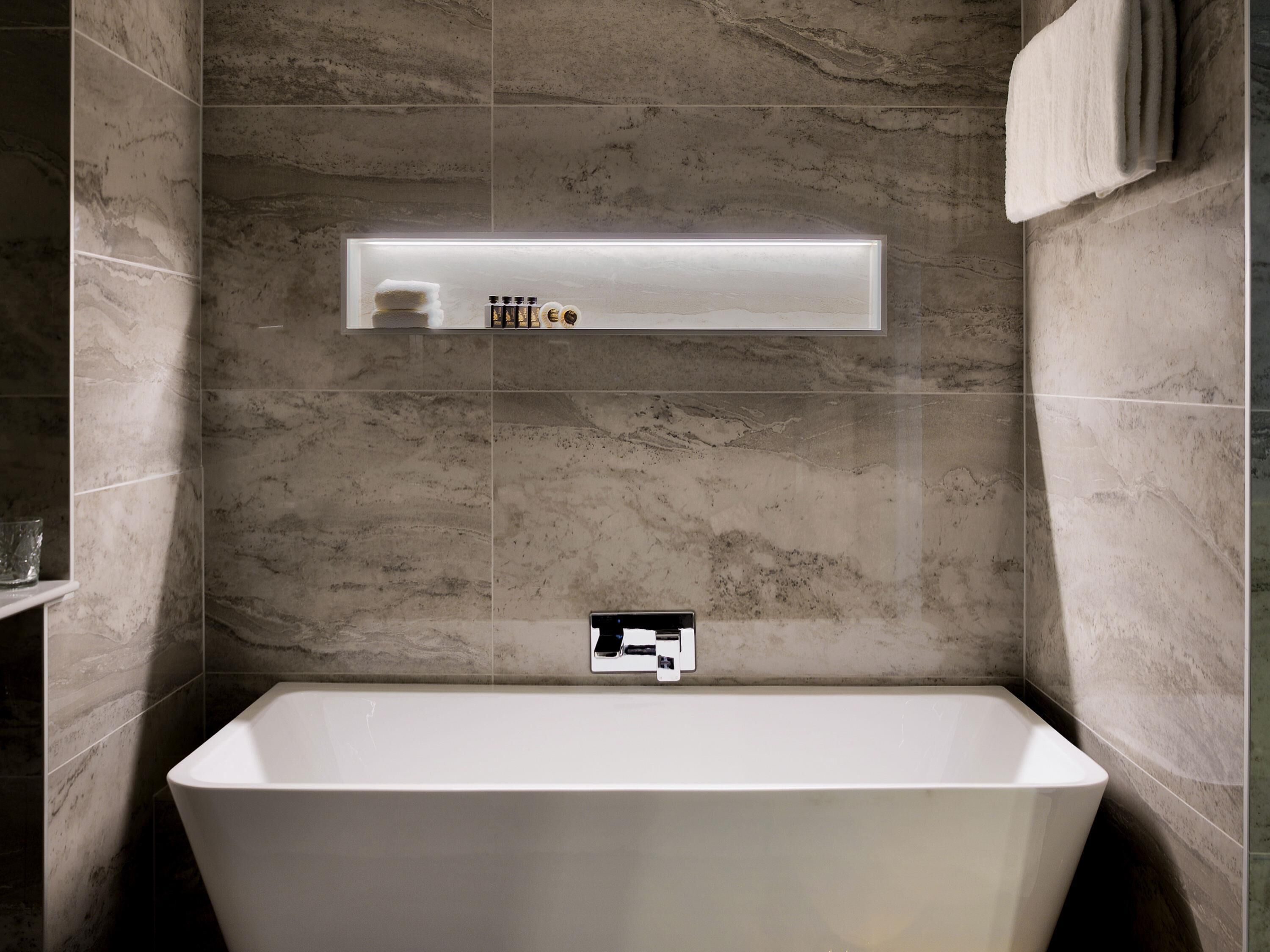 A modern bathroom with a freestanding bathtub, marble walls, and a built-in shelf with toiletries.