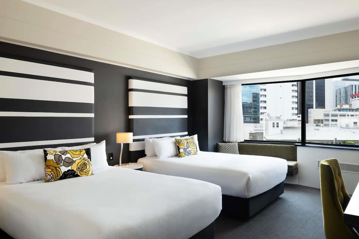 A hotel room with two beds, a window with a city view, a small table, and a chair.