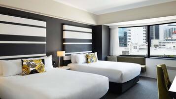 A hotel room with two beds, a window with a city view, a small table, and a chair.