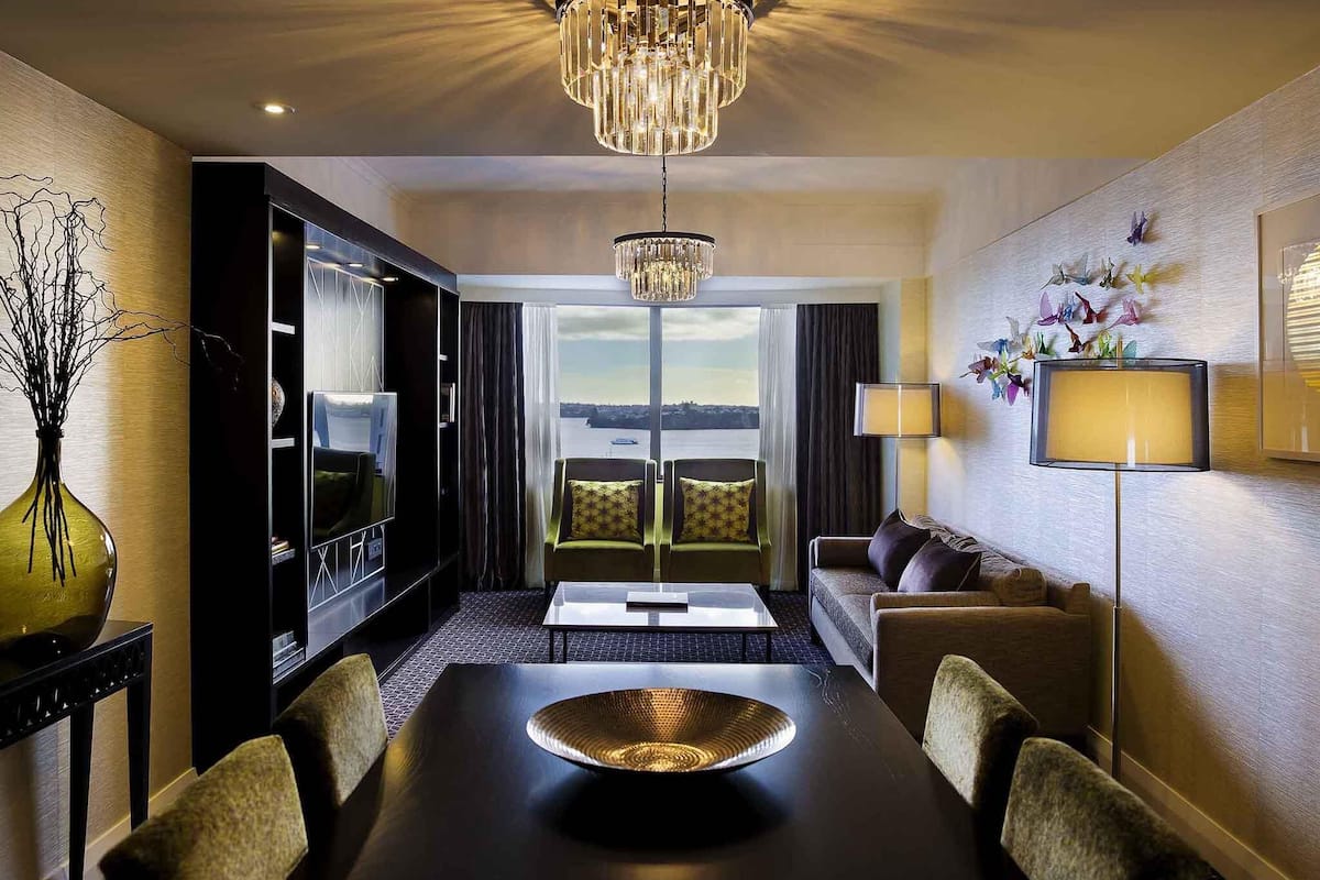 A hotel room with a dining table, chairs, a chandelier, a sofa, a TV, and a view of the city.