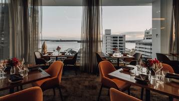 Un restaurant avec des tables dressées, de grandes fenêtres ornées de rideaux et une vue sur un plan d’eau et des bâtiments.