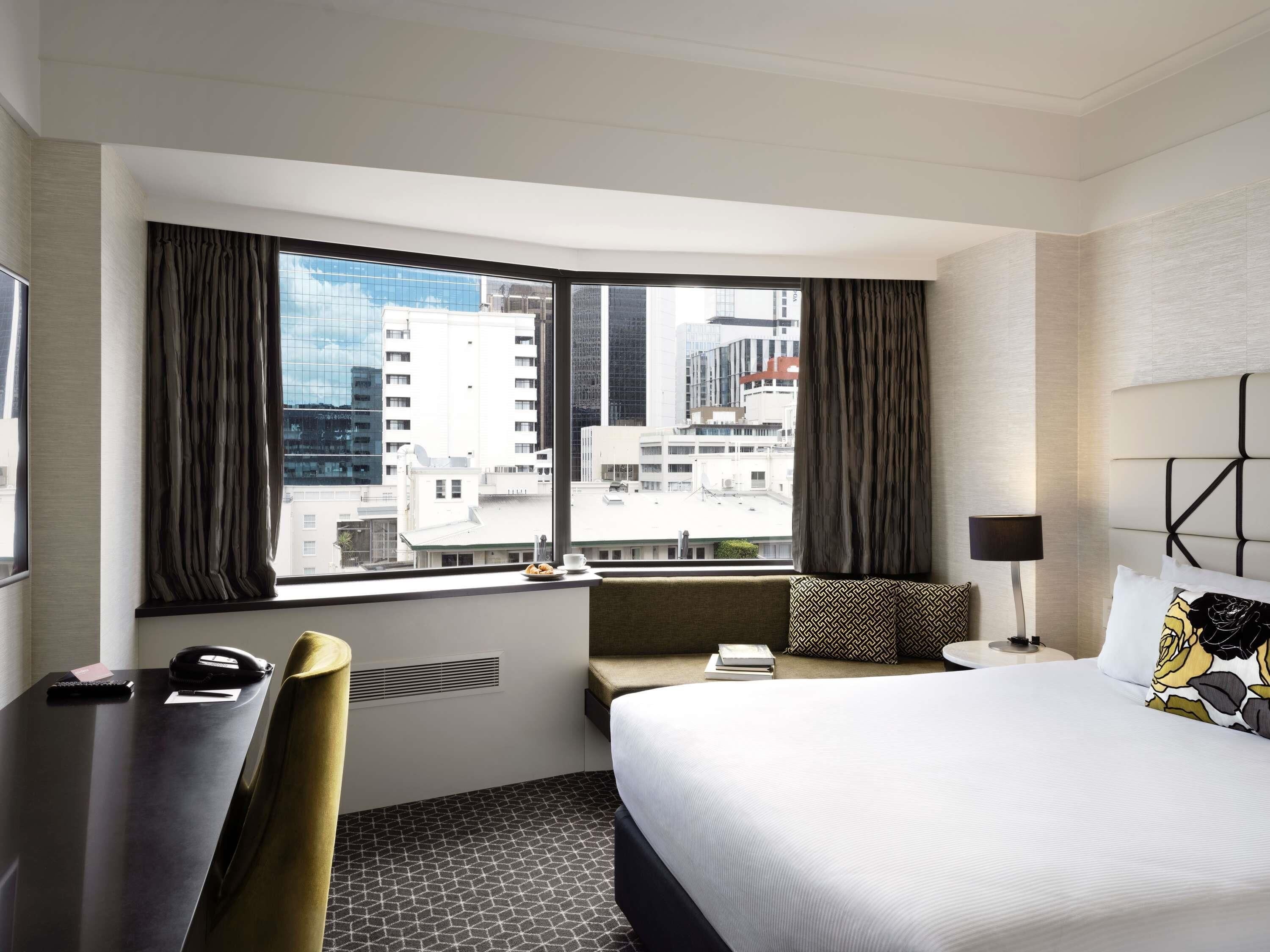 A hotel room with a large bed, a desk, a chair, a bedside table, a lamp, and a view of the cityscape through large windows.