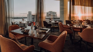 Ein Restaurant mit Blick auf ein modernes Gebäude und ein Gewässer. Tische sind mit Weingläsern und Besteck gedeckt.
