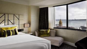 A hotel room with a bed, a chair, a nightstand, a lamp, and a large window with a view of a body of water and a building.