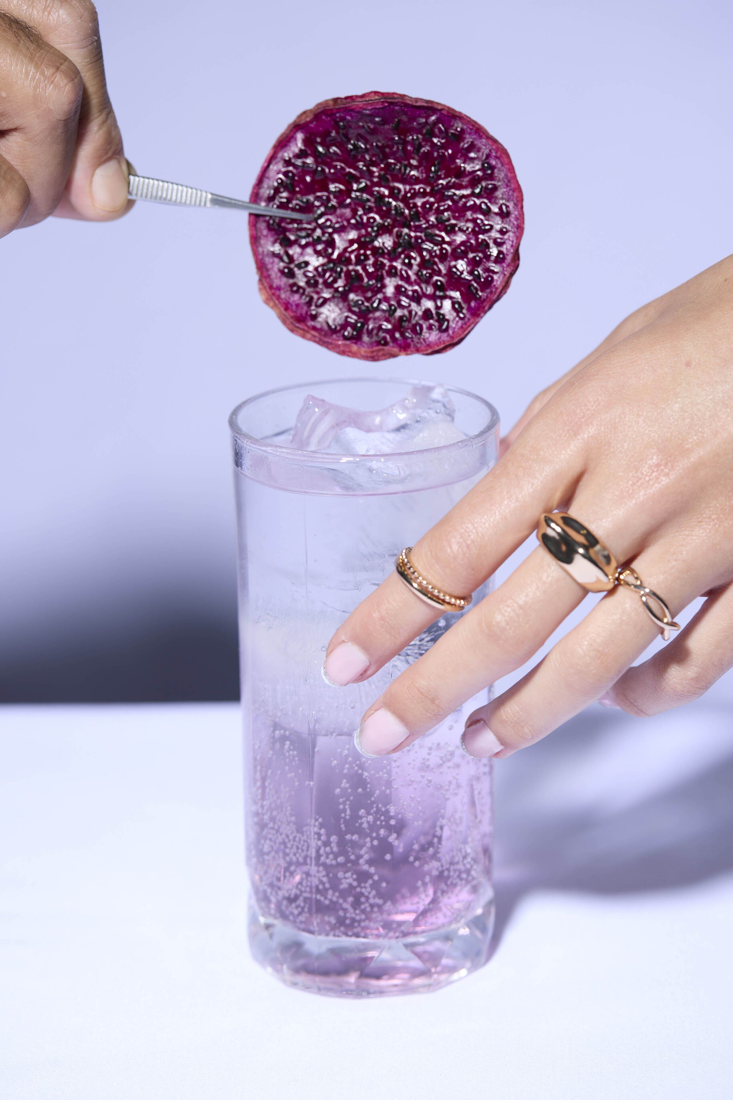 A hand holding a spoon over a glass with ice cubes, about to add a purple fruit.