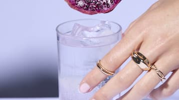 A hand holding a spoon over a glass with ice cubes, about to add a purple fruit.