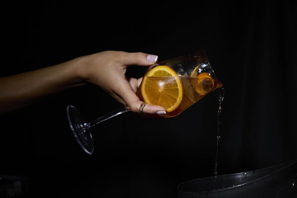 A hand holding a glass with an orange slice and a straw, pouring liquid into a black container.