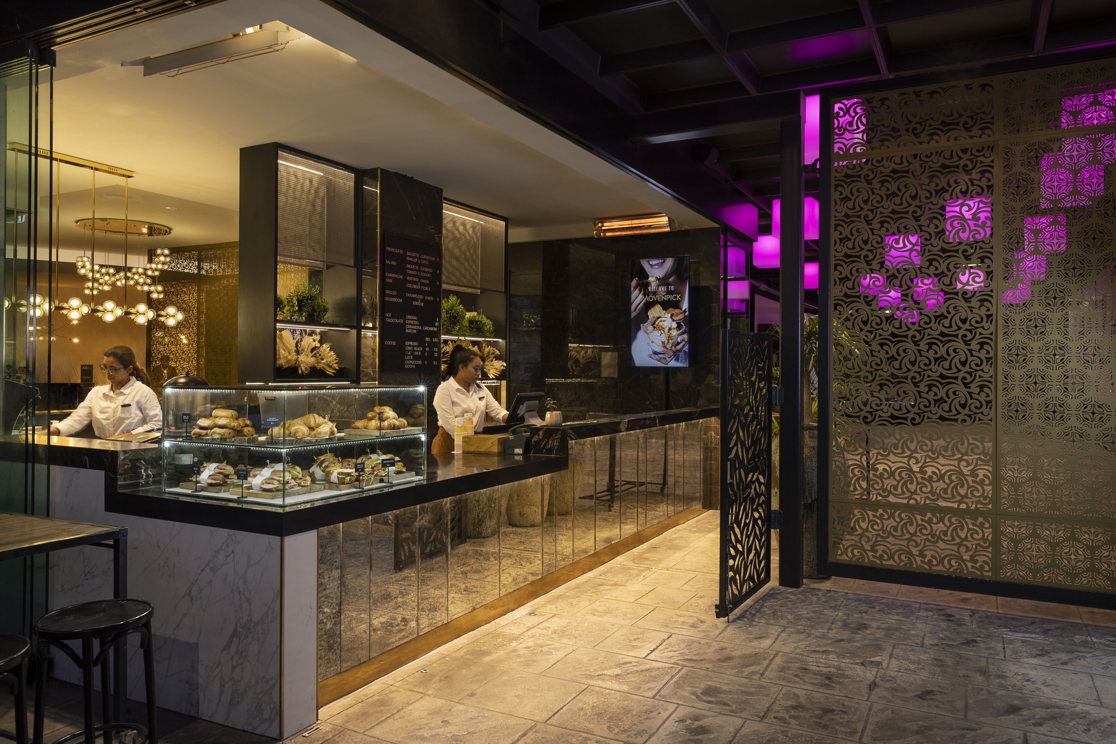 A modern restaurant with a counter displaying food items, two waitstaff, and decorative lighting.