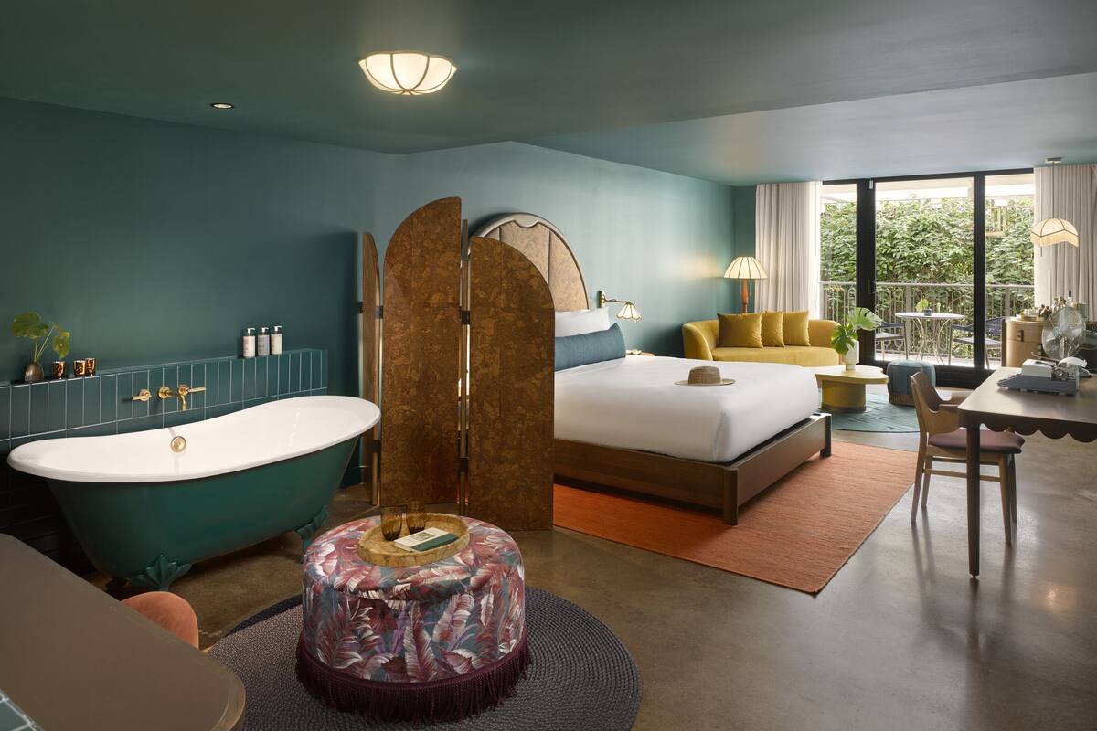 A modern hotel room with a bathtub, a bed with a headboard, a dining area, and a balcony with a view.