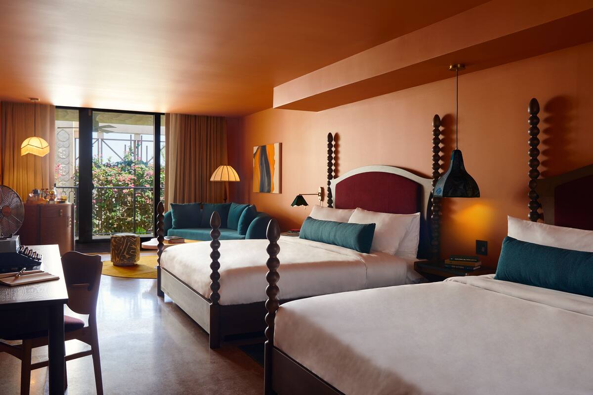 A hotel room with two beds, a desk, a chair, and a window with a view of greenery.