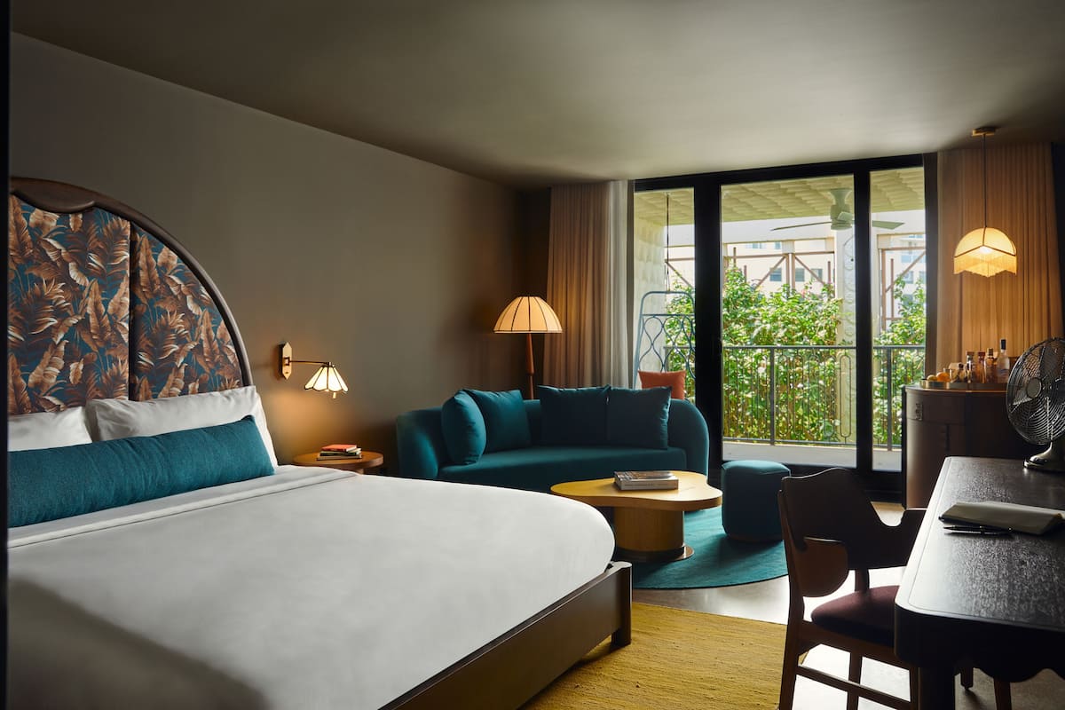A hotel room with a bed, a sofa, a dining table, and a view of greenery outside.