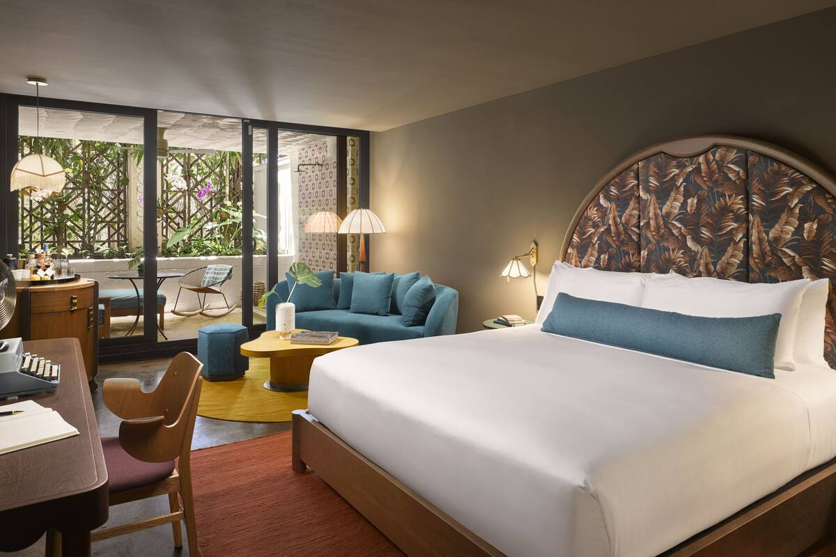 A hotel room with a large bed, a desk with a chair, a sofa, and a small table. There is a window with a view of greenery.