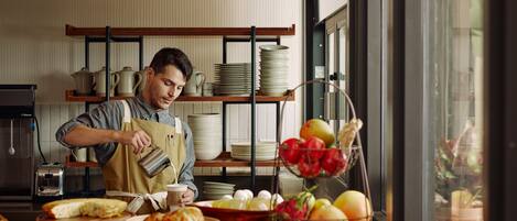 A kitchen with a chef preparing food, a fruit bowl, and shelves with plates and pots.