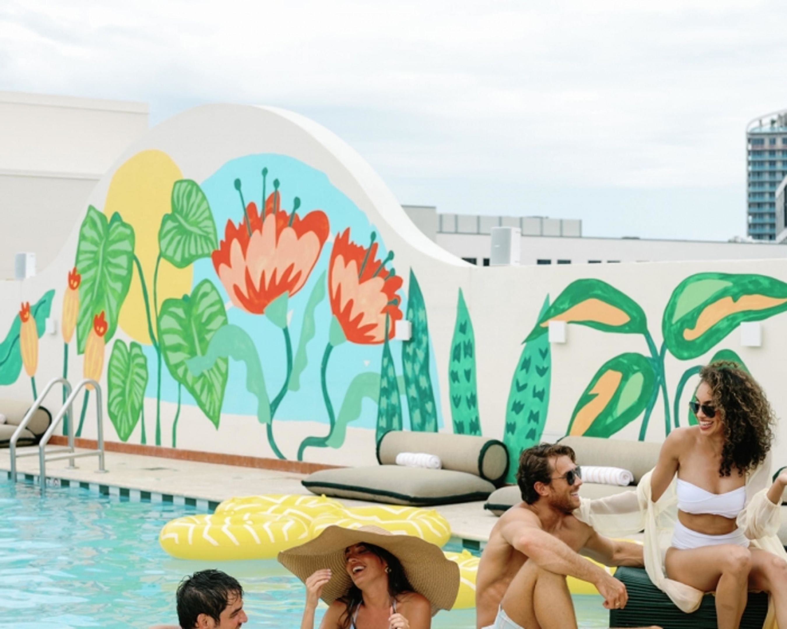 A pool area with a mural of tropical plants and flowers, lounge chairs, and a clear sky.
