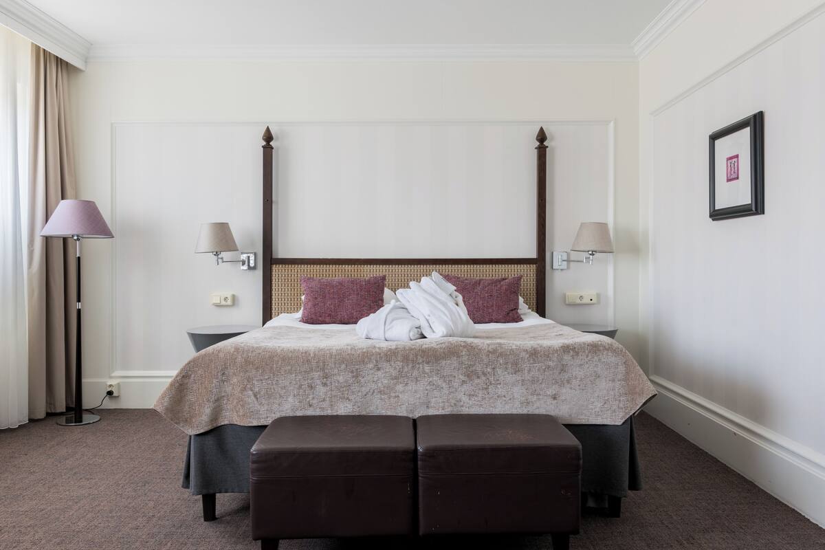 Una habitación de hotel con una cama con dosel, dos lámparas de noche, un ventanal con cortinas, un banco marrón oscuro y un cuadro en la pared.