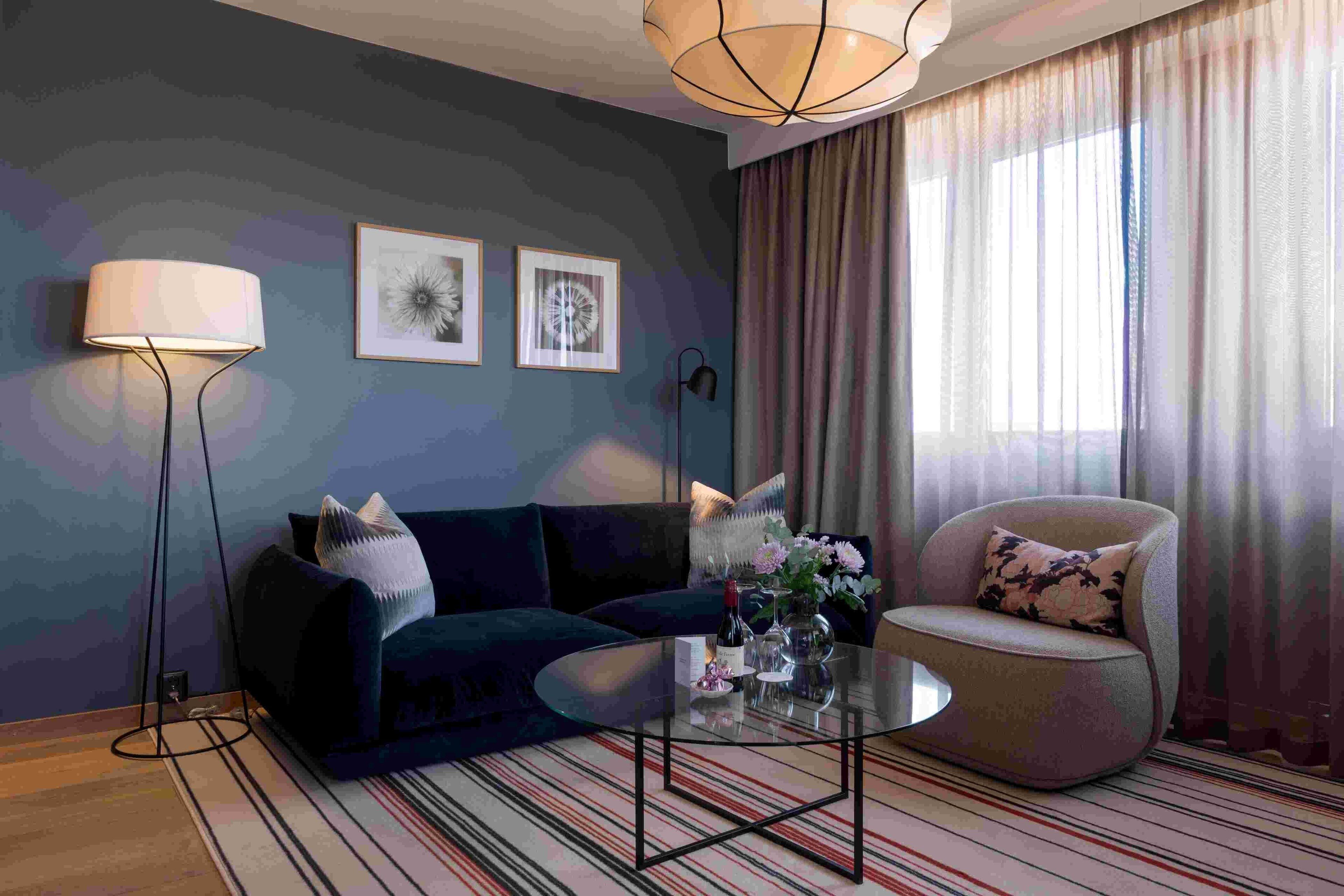 Una sala de estar moderna con un sofá azul oscuro, una mesa redonda de centro y un sillón color beige. Hay obras de arte enmarcadas en la pared, una lámpara de pie y un ventanal con cortinas.