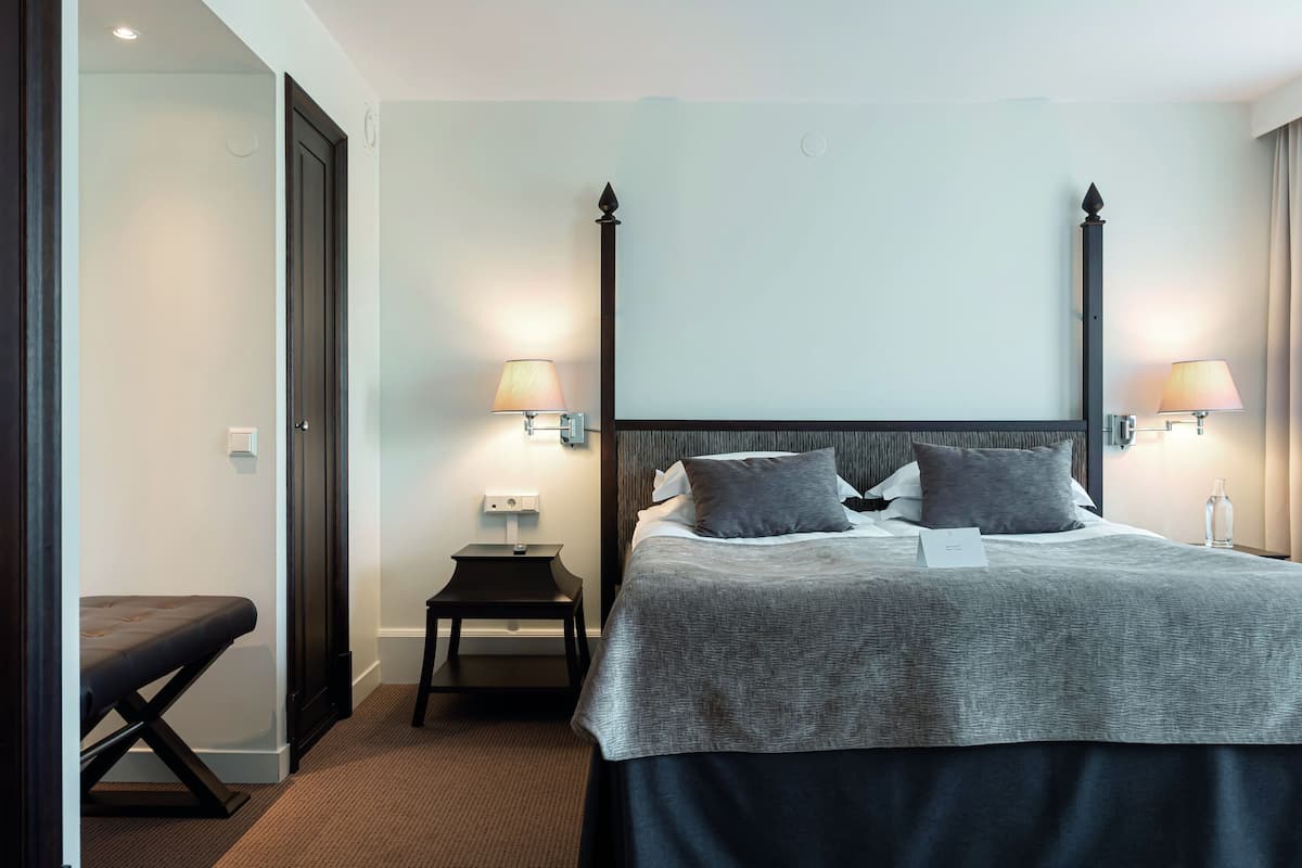 Quarto de hotel bem arrumado, com uma cama grande, abajures, uma mesinha e um banquinho.