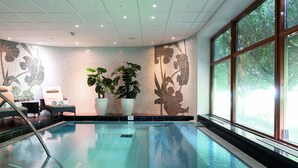 Una piscina interior con agua cristalina, rodeada de grandes ventanales, plantas en macetas y una pared de azulejos con motivos florales.