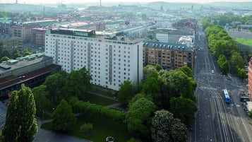 En stor byggnad med flera våningar och en modern arkitektonisk stil, omgiven av grönområden och en livlig gata där en spårvagn passerar.