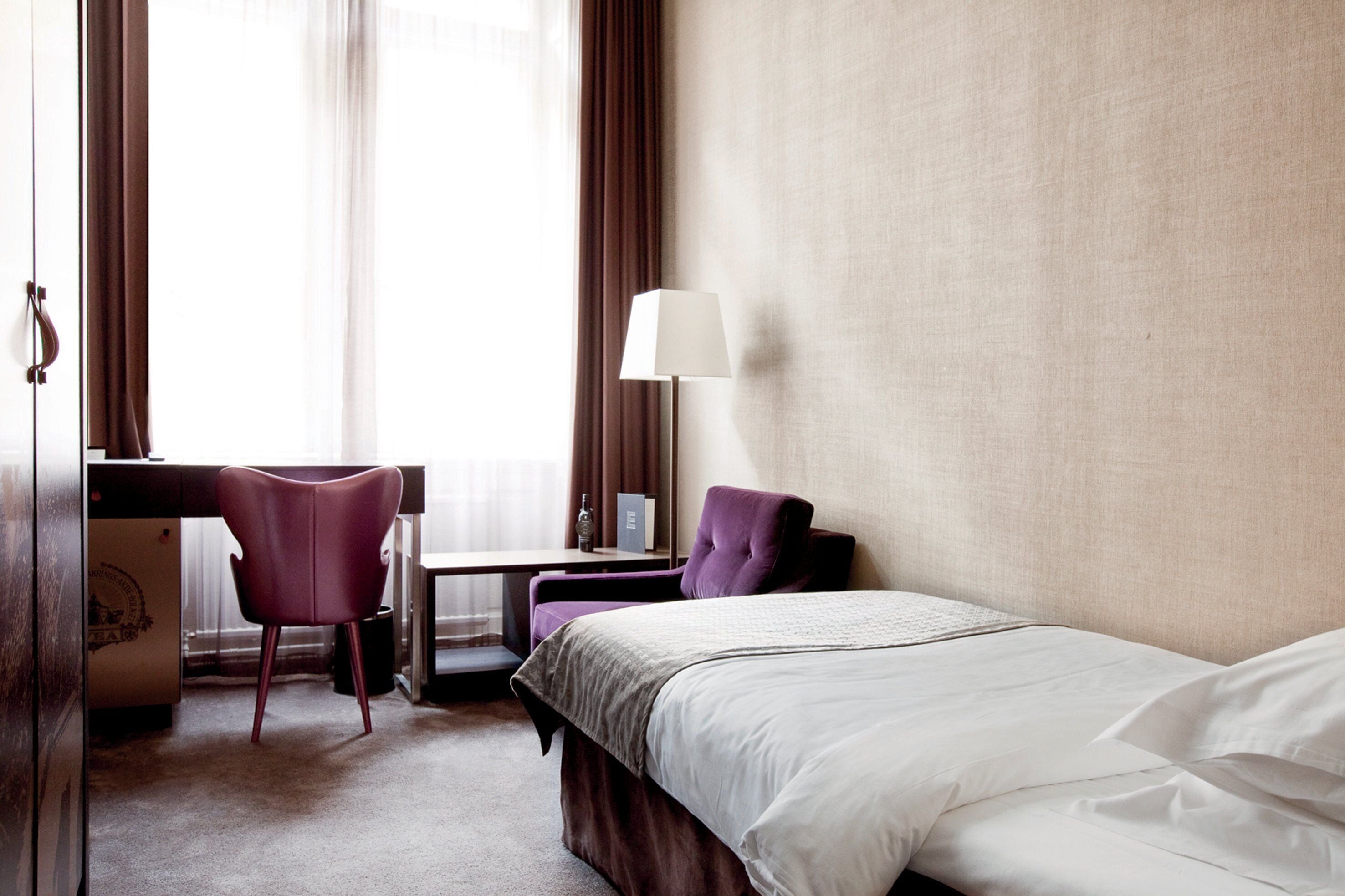 A hotel room with a bed, a desk, a chair, and a lamp.