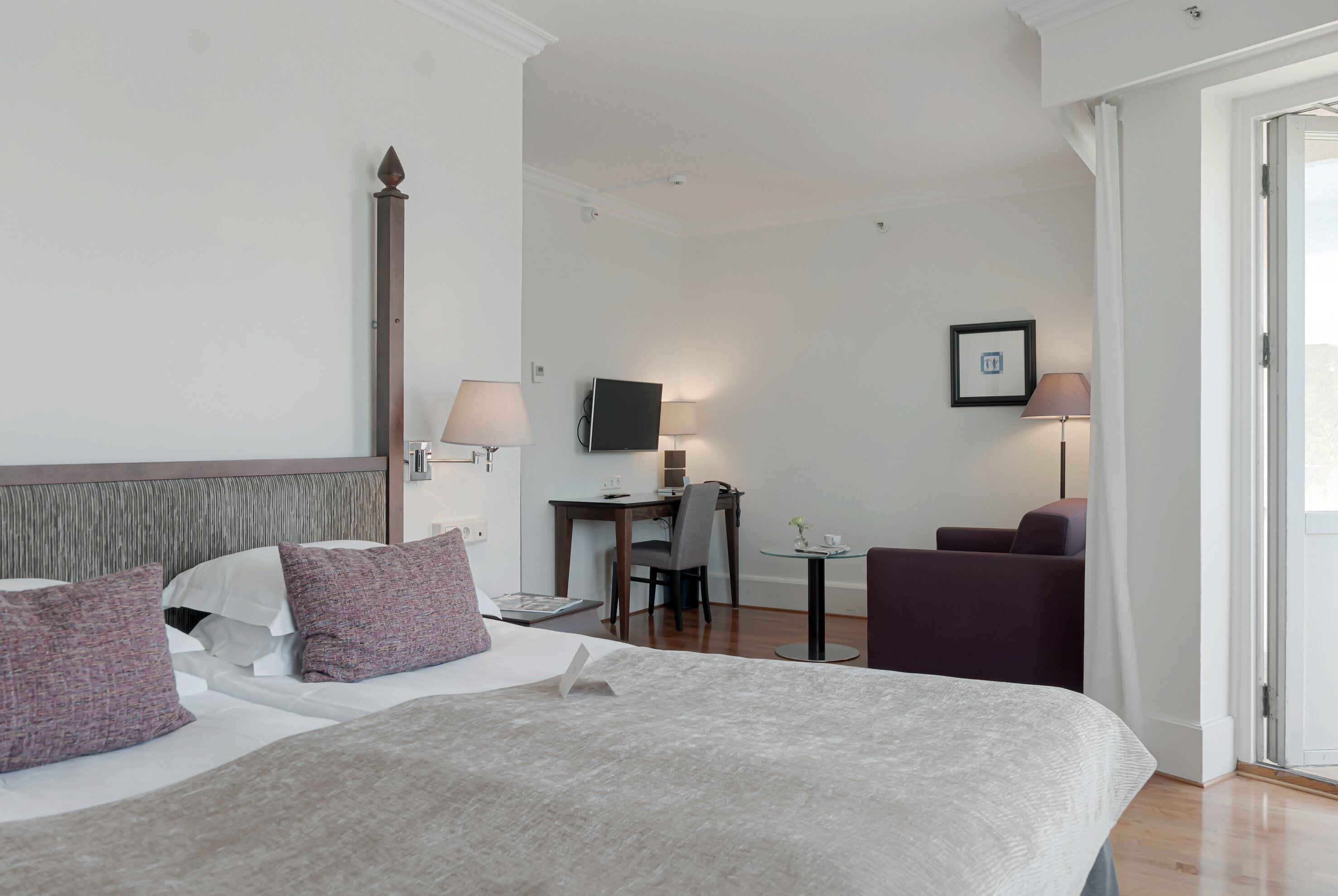 Habitación de hotel con una cama grande, un escritorio con silla, una mesita de noche con lámpara y un sillón púrpura.