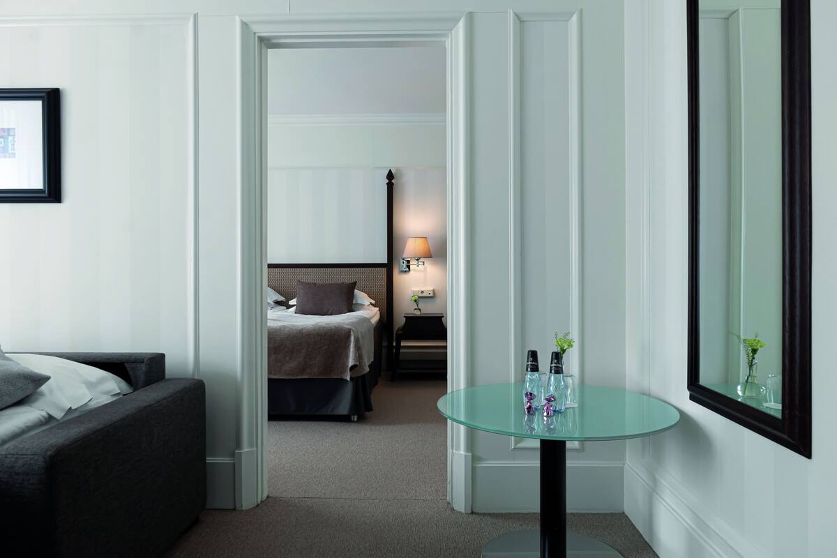 Quarto de hotel com uma cama, mesa de cabeceira com abajur, mesa de vidro com produtos de higiene pessoal e um espelho grande.