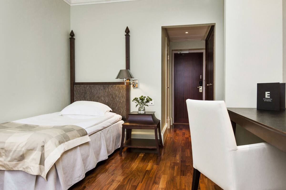 Una habitación de hotel con una cama, una mesita de noche con una lámpara, una silla y una puerta que da a otra habitación.