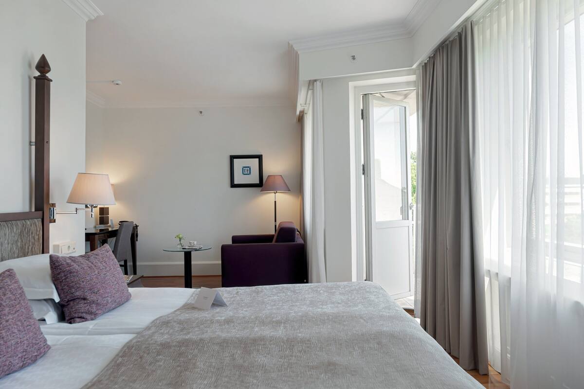 Habitación de hotel con cama, dos mesitas de noche, una silla, un escritorio y ventana con cortinas.