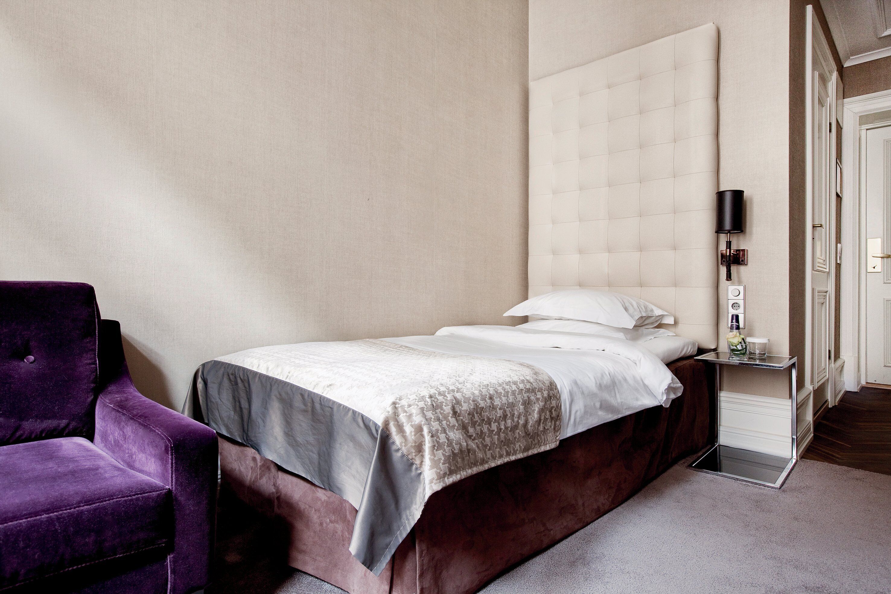 Un dormitorio con una cama, una silla morada, una mesita de noche con lámpara y una puerta.