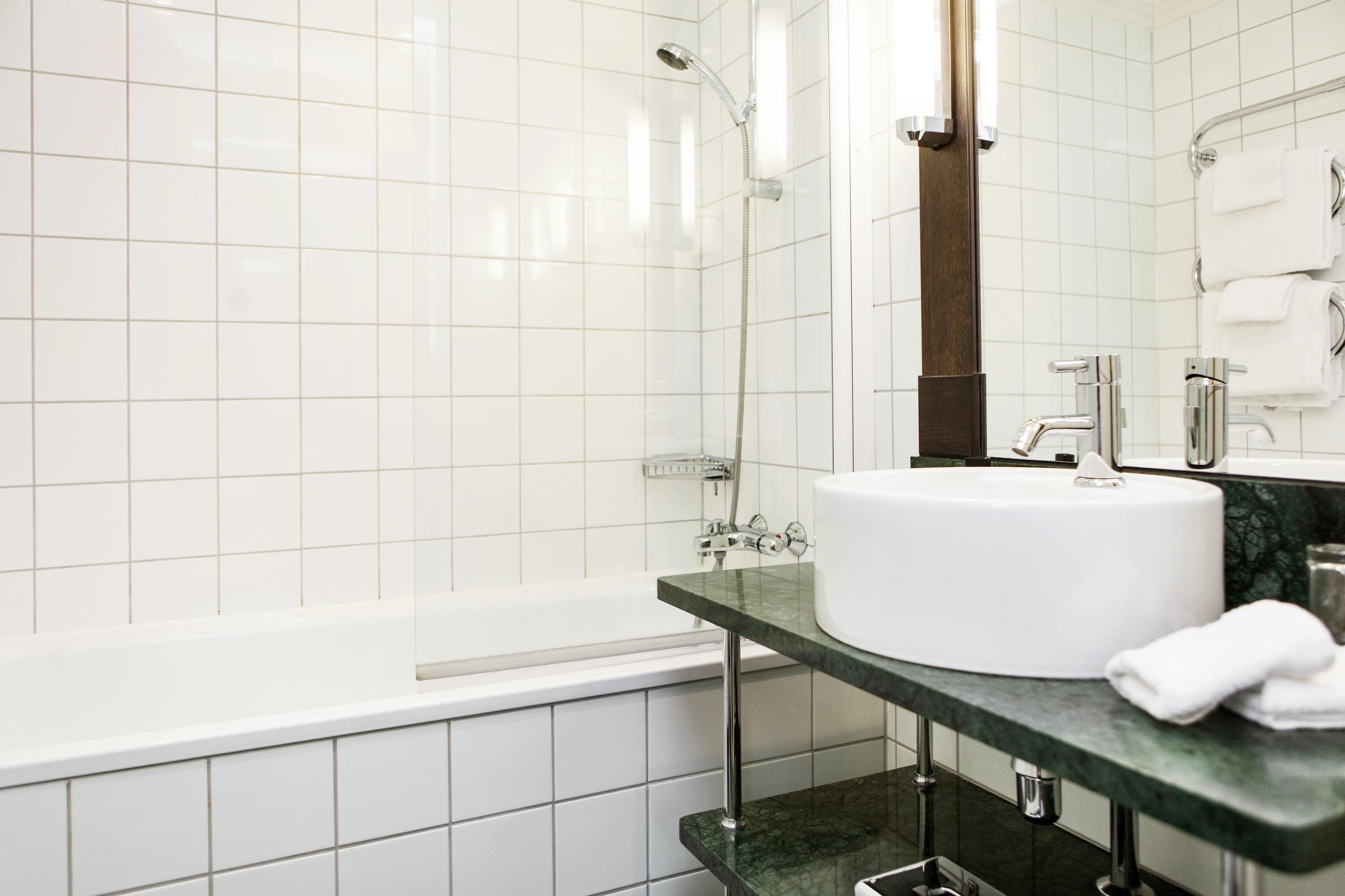 Ett modernt badrum med vita kakelbeklädda väggar, ett vitt badkar, en grön marmorskiva, ett vitt handfat och en stor spegel.