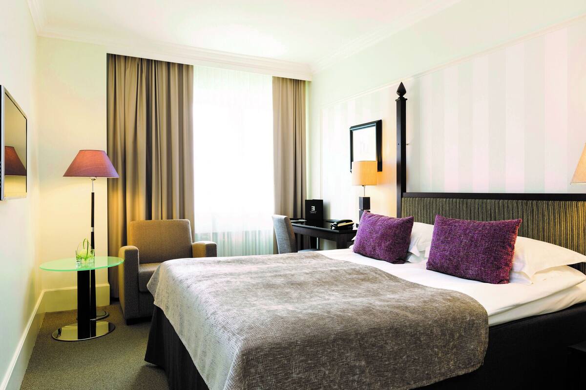 Quarto de hotel com uma cama grande, uma escrivaninha, uma cadeira e uma mesinha de cabeceira com abajur.
