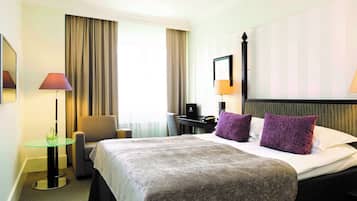 A hotel room with a large bed, a desk, a chair, and a small table with a lamp.
