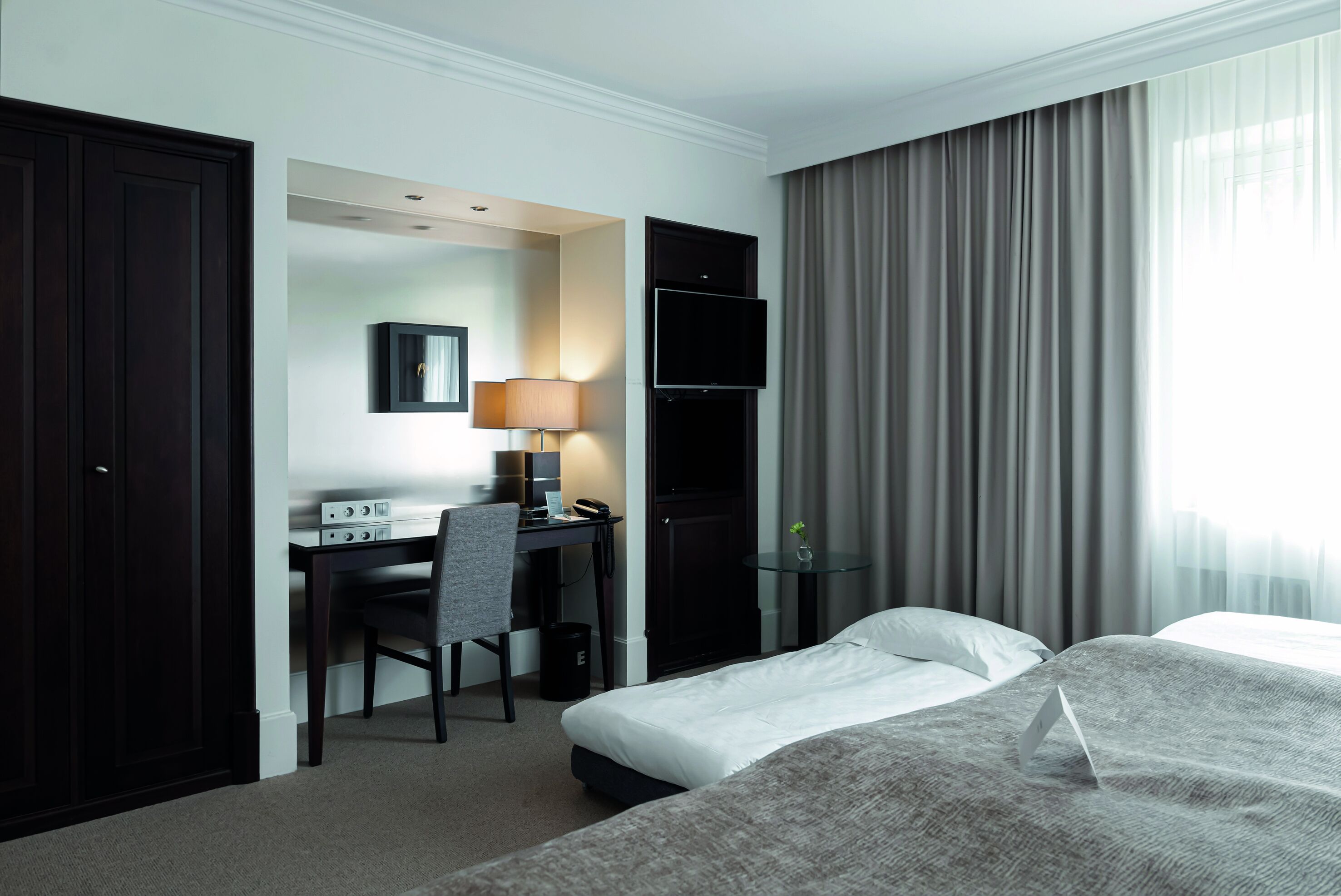 A hotel room with a bed, a desk with a computer, a TV, and a window with curtains.